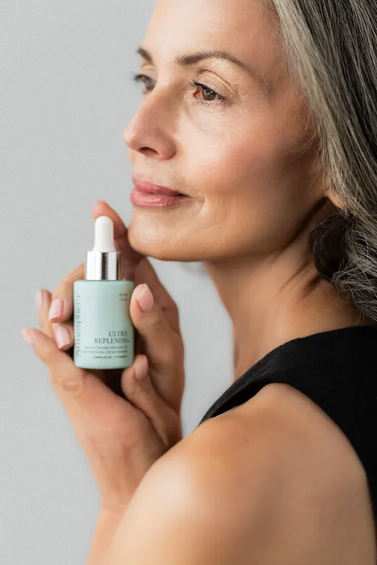 A woman with silver hair holding a bottle of ULTRA REPLENISH™ at her jaw.