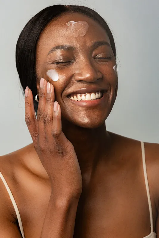 A woman laughing with her eyes closed, dabs of white moisturizer on her forehead and cheeks.
